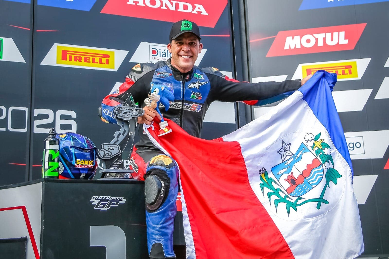 Hilton no pódio do Moto 1000 GP com troféu, Sonic e bandeira de Alagoas