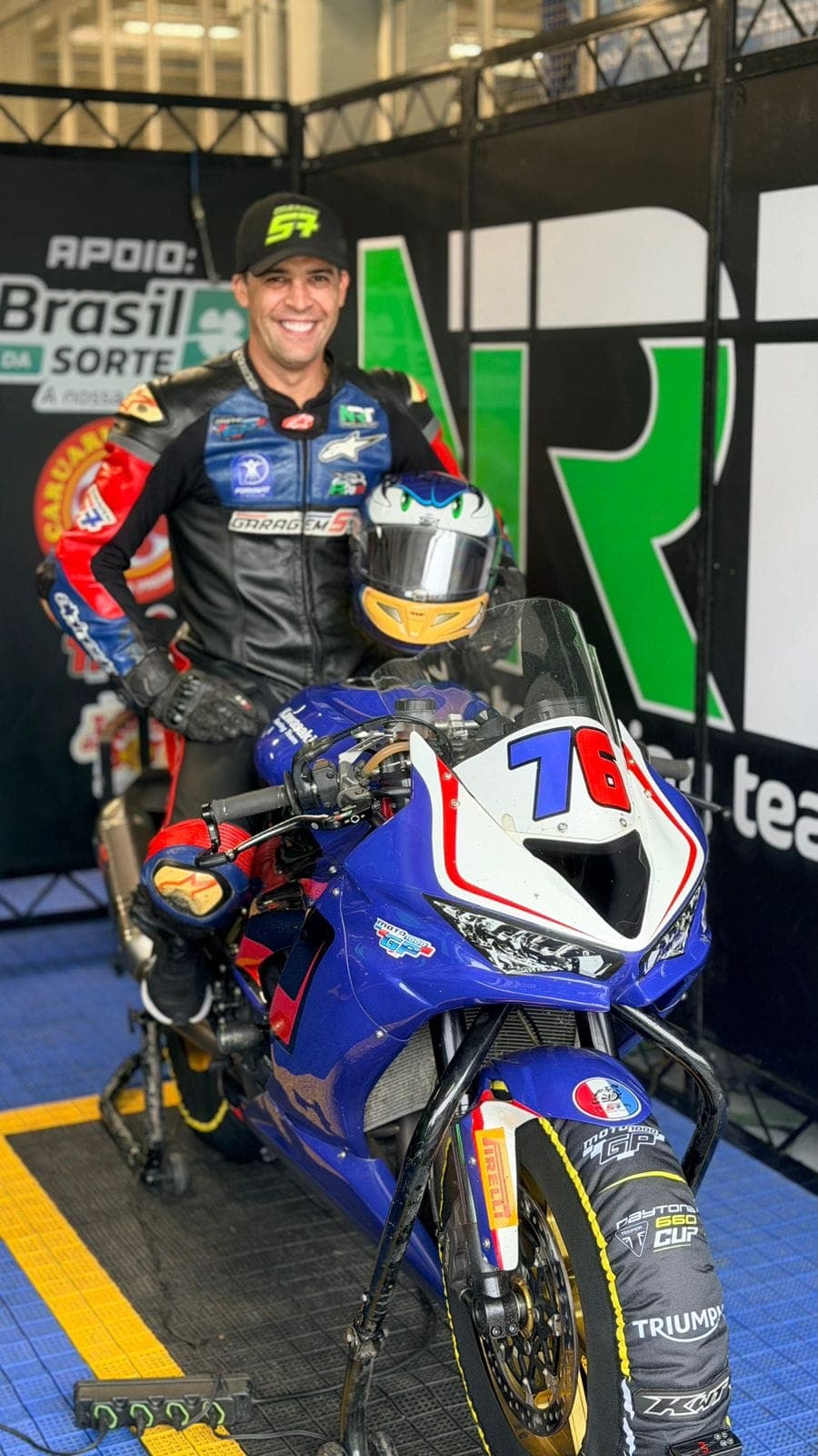 Hilton ao lado da moto #76 no box NRT, segurando o capacete Sonic