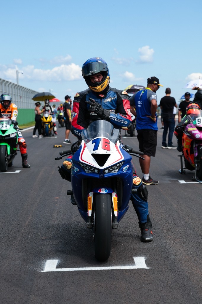 Hilton Loureiro de pé no grid de largada ao lado da Kawasaki ZX6R número 76, capacete azul com adesivo 76, traje vermelho, preto e branco, gesto de positivo com a mão